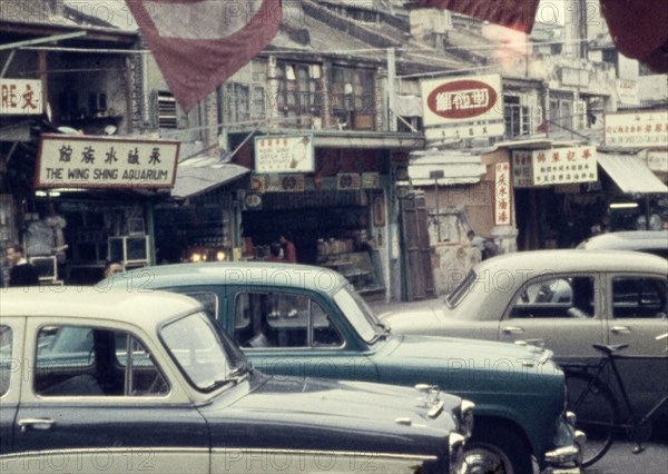Street in Kowloon. A line of cars is parked outside shopfronts on a street in central Kowloon. Kowloon, People's Republic of China, March 1960. Kowloon, Hong Kong, China, People's Republic of, Eastern Asia, Asia.