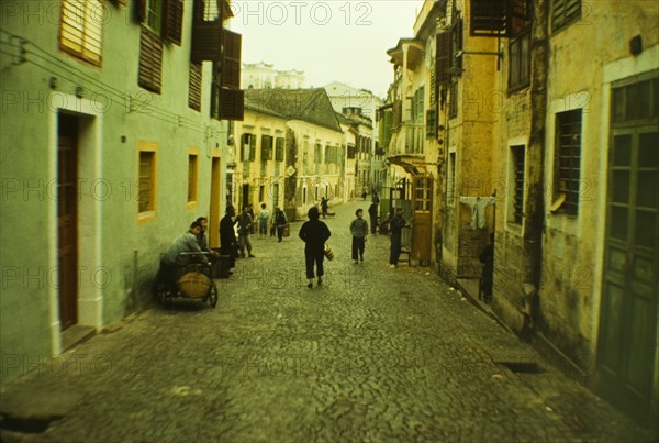 Street scene in Macau. People go about their daily business on a cobbled street bordered by brightly painted houses in the centre of Macau. Macau, People's Republic of China, 2 January 1961. Macau, Macau, China, People's Republic of, Eastern Asia, Asia.