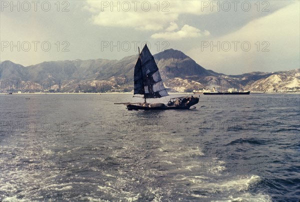 A Chinese junk. A Chinese junk pictured against mountains looking towards Kai Tak. Kai Tak, Hong Kong, People's Republic of China, July 1960. Kai Tak, Hong Kong, China, People's Republic of, Eastern Asia, Asia.