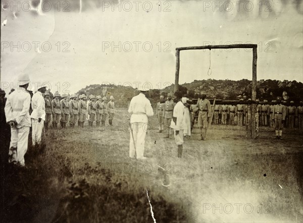 Governor reads death sentence. A European official reads a death sentence, facing a large wooden scaffold with a noose hanging from it. He is surrounded on several sides by uniformed men, both European and African. German East Africa (Tanzania), 1906. Tanzania, Eastern Africa, Africa.
