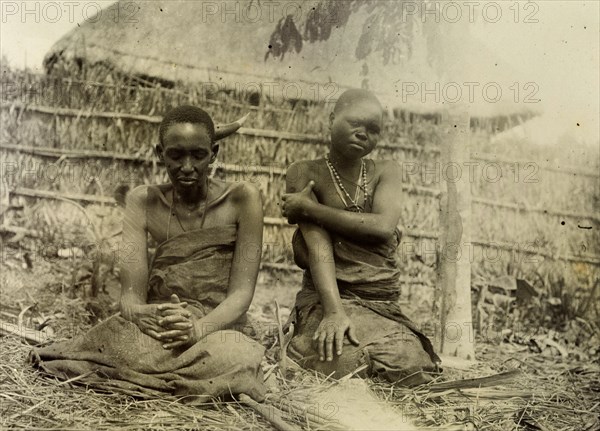 Sufferers from sleeping sickness, Uganda. A man and a woman dressed in bark cloth are seated beneath a tree in a fenced compound. A caption identifies them as sleeping-sickness victims and related photographs suggest that they were part of a government or missionary endeavour to understand and treat the disease. Uganda, 1906. Uganda, Eastern Africa, Africa.