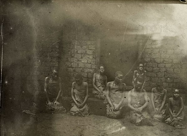 Sufferers from sleeping sickness, Uganda. A group of women and girls suffering from sleeping sickness kneel on the ground beside a doorway, their hands in their laps. One or two of the group can be seen wearing crosses and beads around their necks. Uganda, 1906. Uganda, Eastern Africa, Africa.