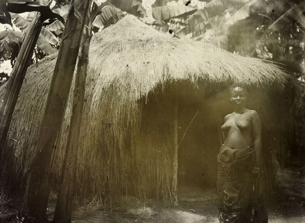 Thatched hut in the forest, Uganda. Portrait of a Ugandan woman standing semi-naked before a thatched village hut in a forest glade. Uganda, 1906. Uganda, Eastern Africa, Africa.