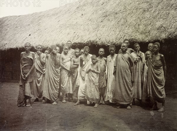 Group portrait, Uganda, 1906. About twenty men, women and children pose for their photograph in front of a large thatched building. All are wearing long garments made of bark cloth or cotton. Uganda, 1906. Uganda, Eastern Africa, Africa.