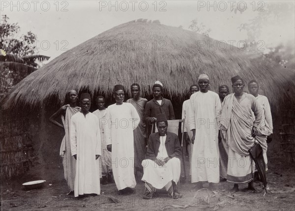 Refugees in Mwanza. A dozen well-dressed men pose for their photograph in front of a low, thatched house. A caption identifies them as 'rebels' who have taken refuge in Mwanza in German East Africa, implying that they have fled there from Uganda. The quality of their dress suggests they are political leaders rather than the foot soldiers of a rebellion. German East Africa (Tanzania), 1906. Tanzania, Eastern Africa, Africa.