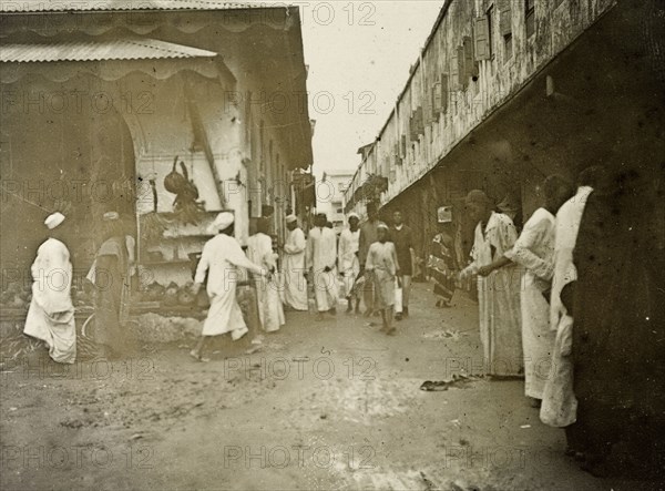 Zanzibar 'business quarter'. Men in long robes walk purposefully along a busy, narrow road in the centre of Zanzibar, originally captioned as the 'business quarter'. A shop on the left can be seen selling fruit and vegetables. Zanzibar (Tanzania), 1906. Zanzibar Town, Zanzibar Urban/West, Tanzania, Eastern Africa, Africa.