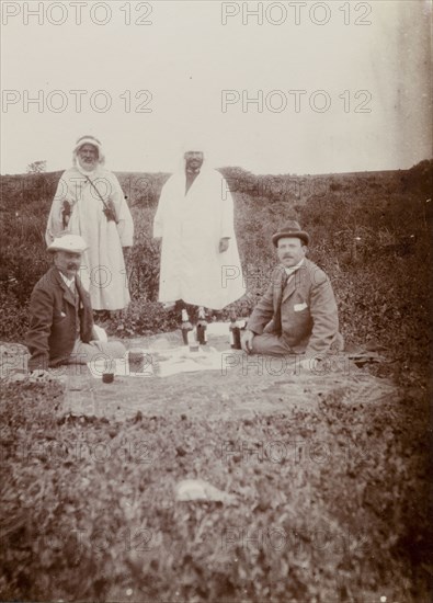 Picnic lunch, Morocco. Two European men enjoy a picnic on a blanket in an open field: their Moroccan guides standing behind them. The European man on the right is Frederick Stanbury, the other, his travelling companion in Morocco, 'Hunter'. The photograph was taken on an excursion to archaeological ruins. Morocco, 1898. Morocco, Northern Africa, Africa.