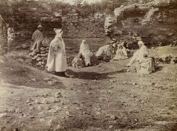 Morrocan men at a ruin. A group of Moroccan men dressed in white robes gather beside a ruin in the desert. A caption suggests that Frederick Stanbury visited these people on an excursion during his travels in Morocco. Morocco, 1898. Morocco, Northern Africa, Africa.