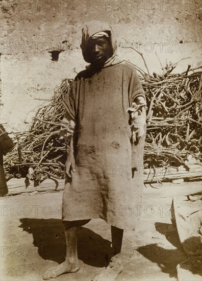 Labourer in Safi, Morocco. Outdoors portrait of a barefoot labourer in ragged clothing, standing in front of a pile of wood. A caption identifies him as a 'worker'. Safi, Morocco, 1898. Safi, Safi, Morocco, Northern Africa, Africa.
