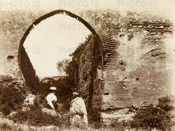 Islamic-style arch, Morocco. A Moroccan man talks to a Europan woman beneath an Islamic-style brick arch at a ruined fort. Southern Morocco, 1898. Morocco, Northern Africa, Africa.