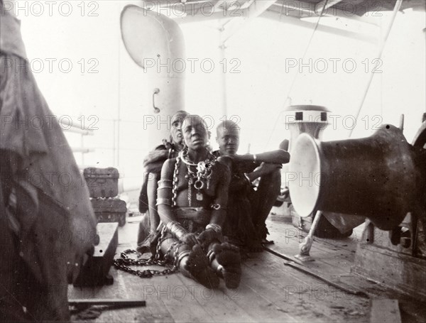 African woman convict on steamer deck. A semi-naked African woman sits on the deck of a steamer on Lake Victoria, bound by heavy chains. The two African men sitting behind her may be her guards. A caption identifies her as a 'poisoner' who is being taken to Muanza (Mwanza) in German East Africa (Tanzania) for punishment. German East Africa (Tanzania), 1906., Kagera, Tanzania, Eastern Africa, Africa.