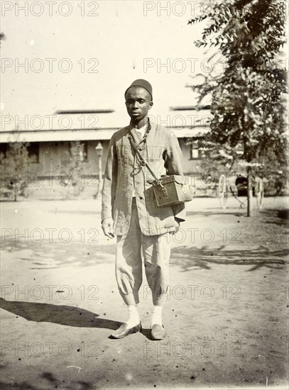 Frederick Stanbury's domestic servant. Stanbury's servant is an unidentified young African man. He stands outside a colonial bungalow wearing a Western-style jacket and short trousers, shoes, socks and a fez. A square leather box is slung around his neck and he holds a cigarette. British East Africa (Kenya), 1906. Kenya, Eastern Africa, Africa.