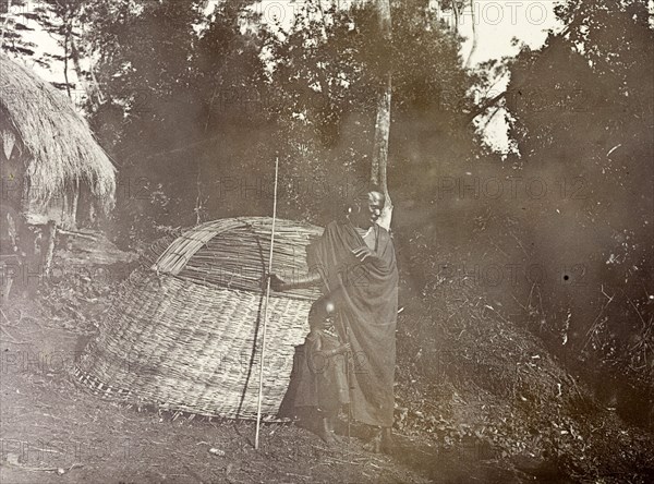 Wickerwork village hut. A Kenyan man and child stand are pictured in their village beside a partially-completed wickerwork hut. British East Africa (Kenya), 1906. Kenya, Eastern Africa, Africa.