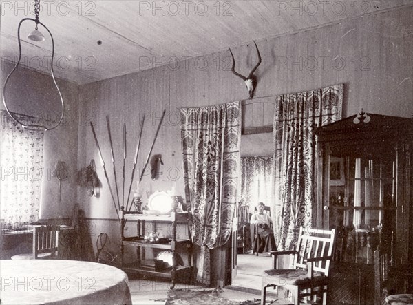 Mrs H' in the drawing room. A European woman identified as 'Mrs H', is seated in her drawing room, seen through partly drawn curtains. The room in the foreground displays a variety of cross-cultural objects including African spears and a Western-style tea service. British East Africa (Kenya), 1906. Kenya, Eastern Africa, Africa.