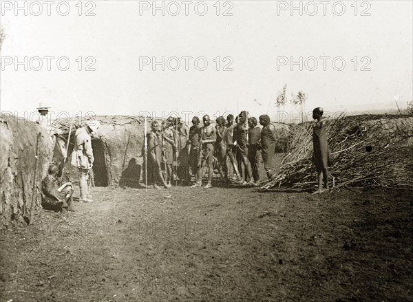Inside a Maasai village. A group of Maasai watch as two European men examine a wall made from mud inside their village. British East Africa (Kenya), 1906. Kenya, Eastern Africa, Africa.