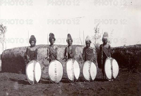 Maasai warriors. Maasai society is ordered by age groups and the five men shown here are either junior or senior warriors. They wear lion's mane headdresses and hold shields made of toughened hide. Behind them can be seen the typical low, breadloaf-shaped house of Maasai communities. British East Africa (Kenya), 1906. Kenya, Eastern Africa, Africa.