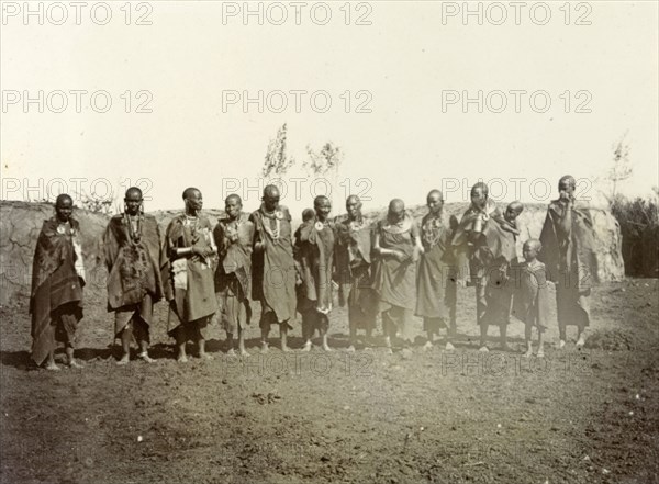 Maasai adults and children. A group of Maasai adults and children wearing traditional dress and jewellery line up for the camera. Behind them can be seen the low, breadloaf-shaped houses typical of Maasai communities. British East Africa (Kenya), 1906. Kenya, Eastern Africa, Africa.