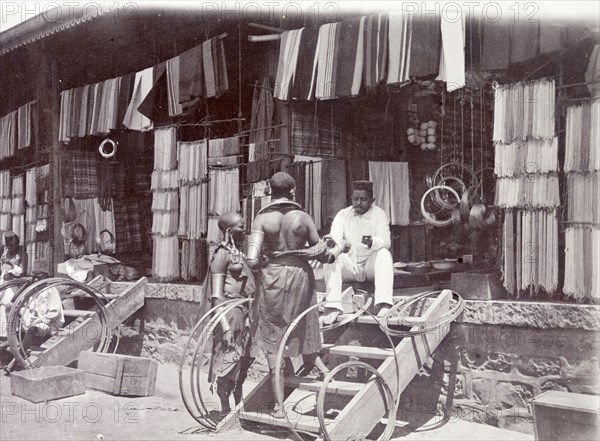 Indian shop, Nairobi. An Indian trader sits in the open doorway of his store, chatting to two African customers. Nairobi, British East Africa (Kenya), 1906. Kenya, Eastern Africa, Africa.