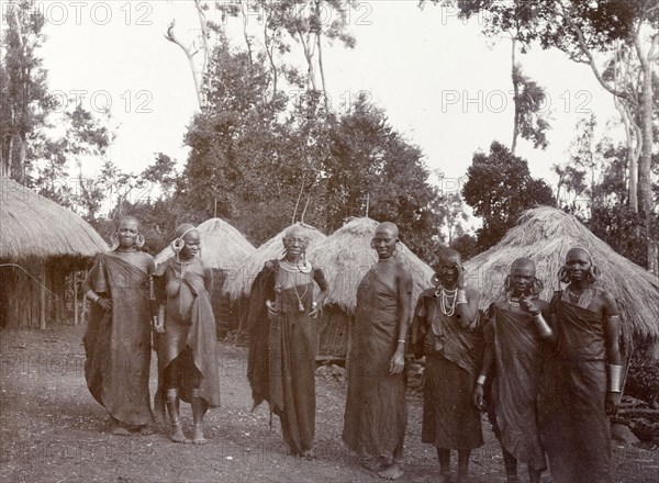 Kikuyu women in a village. Seven Kikuyu women pose for their photograph in front of their round, grass-roofed houses. British East Africa (Kenya), 1906. Kenya, Eastern Africa, Africa.