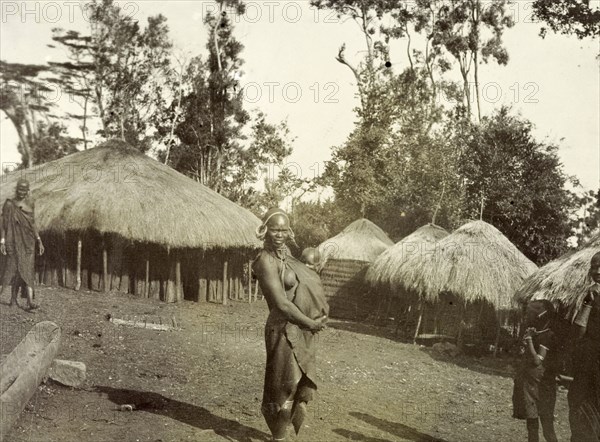 Domestic village scene, Kikuyu. Domestic village scene. A Kikuyu woman wearing traditional dress and masses of earrings, holds a baby close to her body. Kikuyu, British East Africa (Kenya), 1906. Kikuyu, Central (Kenya), Kenya, Eastern Africa, Africa.