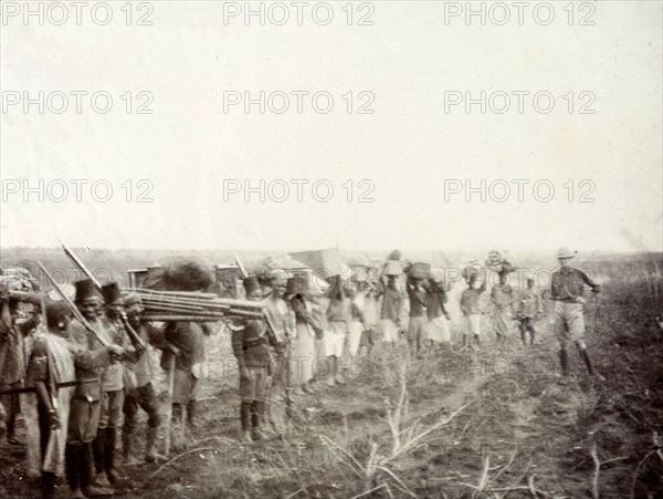 Frederick Stanbury bids farewell. A group of African porters and servants wait in line holding various items of luggage as Frederick Stanbury bids farewell to Geoffrey Archer (pictured), his travelling companion on safari. British East Africa (Kenya), 1906. Kenya, Eastern Africa, Africa.