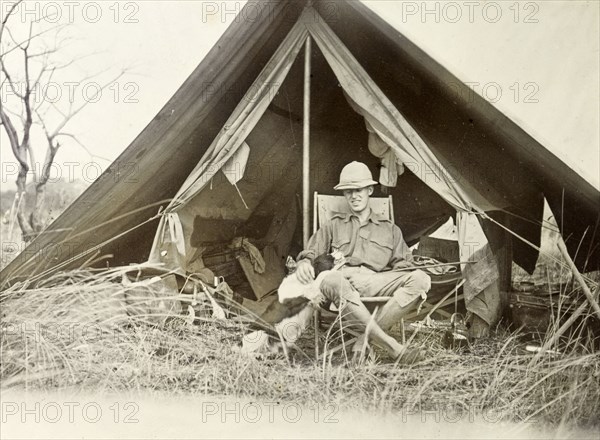 Geoffrey Archer outside a tent. Colonial officer, Geoffrey Archer, is seated on a folding chair in front of a small open tent, a pet dog by his side. Near Lake Baringo, British East Africa (Kenya), 1906. East Africa Protectorate (Kenya), 1906., Rift Valley, Kenya, Eastern Africa, Africa.