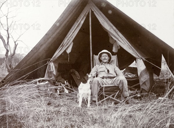 Frederick Stanbury outside a tent. Stanbury is seated on a canvas chair in front of a small open tent, a pet dog by his side. Near Lake Baringo, British East Africa (Kenya), 1906., Rift Valley, Kenya, Eastern Africa, Africa.
