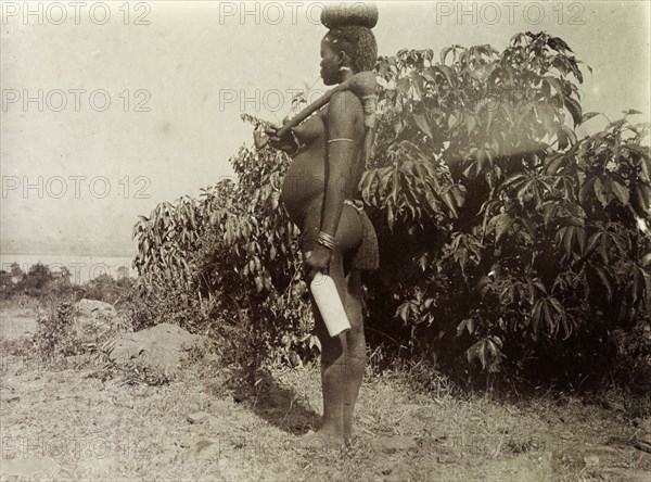 Kavirondo woman. Profile shot of a naked Kenyan woman carrying a bottle of milk on her way to a local market. She rests a hand tool over her shoulder and balances another pot on her head. Kavirondo, British East Africa (Kenya), 1906. Kavirondo, Nyanza, Kenya, Eastern Africa, Africa.