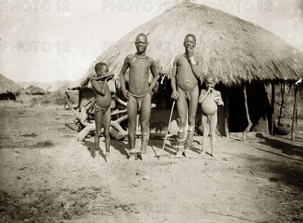 Luo men and children. Two nearly-naked men and two children pose for the camera outside a round, grass-roofed dwelling. Near Kisumu, British East Africa (Kenya), 1906., Nyanza, Kenya, Eastern Africa, Africa.