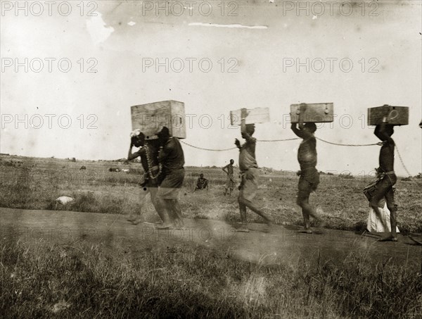 African convicts chained by the neck. A line of African convict porters carry crates on their head or shoulders. They are chained together by the neck and are naked but for a loin cloth. German East Africa (Tanzania), 1906. Tanzania, Eastern Africa, Africa.