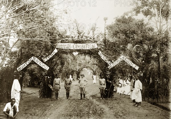 Entrance to the Uganda Exhibition. The entrance to the Uganda Exhibition, fashioned from tied palm leaves and adorned with banners, is guarded by askaris (soldiers). Uganda, circa December 1936. Uganda, Eastern Africa, Africa.