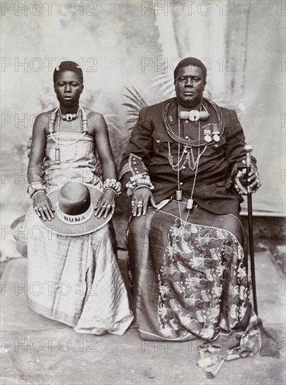 Studio portrait of Dogho Numa and his wife. Studio portrait of Dogho Numa and his wife dressed in formal attire. She wears a long dress with ornate jewellery and holds a wide-brimmed hat decorated with her husband's name 'NUMA'. He wears a military-style jacket decorated with medals over a long wrap-around skirt and carries a staff. Nigeria, circa 1925. Nigeria, Western Africa, Africa.