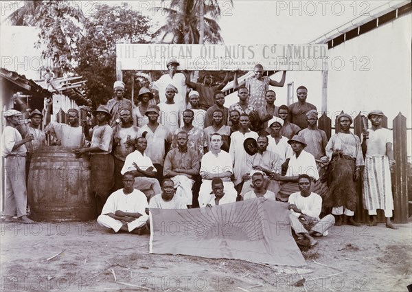 Staff of the African Oil Nuts Company. Group portrait of a European overseer and African staff of the African Oil Nuts Company sitting beneath the company sign. Many of the sitters carry the tools of their trade, several miming actions with them. The five men in the foreground display a large flag, which shows the rising sun and the letters 'AON'. Badagry, Nigeria, May 1922. Badagry, Lagos, Nigeria, Western Africa, Africa.