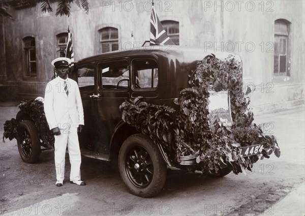 Remembrance Day Car. An African chauffeur stands beside a car decorated with foliage and Union flags for Remembrance Day. A poster on the back of the car exhorts people to wear a poppy for Remembrance Day. Nigeria, 11 November circa 1925. Nigeria, Western Africa, Africa.