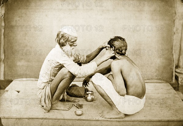 An Indian barber. An Indian barber crouches down on an outdoor platform as he shaves a customer's face. Probably Bengal, India, circa 1900. India, Southern Asia, Asia.