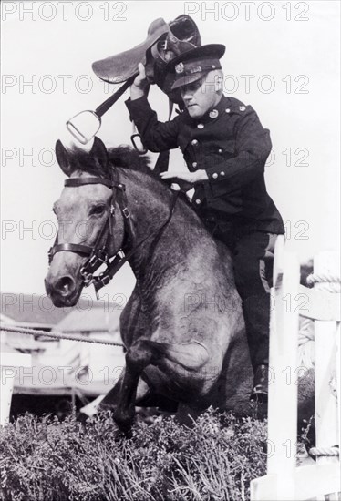 Leslie Slater holds his breath. Uniformed British police officer, Leslie Slater, holds his breath as he jumps over a hedge on horseback, a saddle lifted above his head. This jump was part of a mounted police tournament being held at the Eve Leary parade ground. Georgetown, Guyana, 1948. Georgetown, Demerara-Mahaica, Guyana, South America, South America .