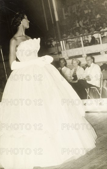 Carnival Queen beauty pageant. A contestant in a beauty pageant turns to smile at a panel of judges as she walks down a catwalk wearing a sleeveless, formal dress and elbow-length gloves. Grenada, 2 March 1957. Grenada, Caribbean, North America .