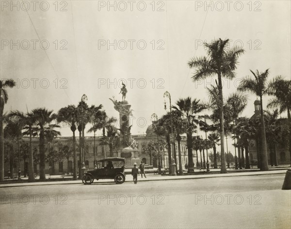 Monument in a piazza, Lima. Palm trees surround a tall stone monument decorated with sculptures in a city piazza. Lima, Peru, circa 1920. Lima, Lima Metropolitana, Peru, South America, South America .