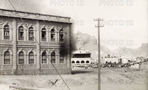 Building set on fire during the Arab riots in Aden, 1947. Black smoke billows from a building in the Jewish quarter of Aden, while crowds of people can be seen milling about at a road junction, during the Arab riots that took place between 1-3 December 1947 in response to the United Nations Partition Plan for Palestine. Aden, Yemen, December 1947. Aden, Adan, Yemen, Middle East, Asia.