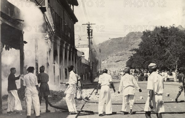Relief work after the Arab riots in Aden, 1947. Teams of men use two hose pipes to douse the flames of a building that has been set on fire in Aden, during the Arab riots that took place between 1-3 December 1947 in response to the United Nations Partition Plan for Palestine. Aden, Yemen, December 1947. Aden, Adan, Yemen, Middle East, Asia.