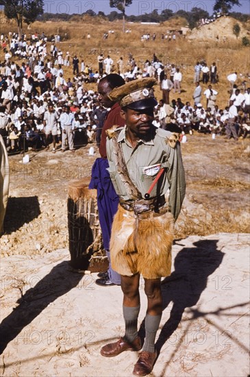 Policeman at United National Indepence Party rally. A policeman on watch at a United National Independence Party (UNIP) political rally. He wears animal fur shorts, with a snakeskin belt and sash, and a snakeskin-trimmed hat on which a badge of UNIP President Kenneth Kaunda is pinned. Kitwe, Northerm Rhodesia (Zambia), circa 1962. Nkana-Kitwe, Copperbelt, Zambia, Southern Africa, Africa.