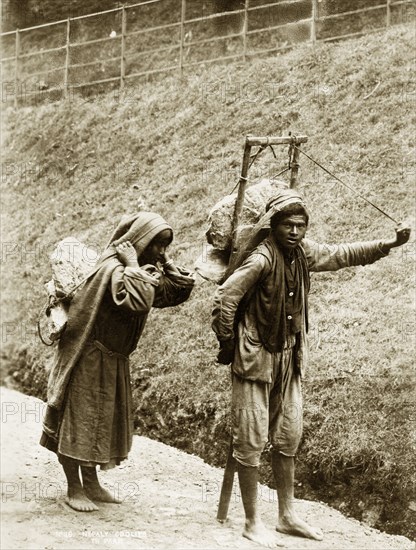 Nepalese labourers. A barefoot Nepalese couple, labelled in an original caption as 'coolies', struggle down a hillside road burdened by large rocks strapped to their backs. Nepal, circa 1890. Nepal, Southern Asia, Asia.