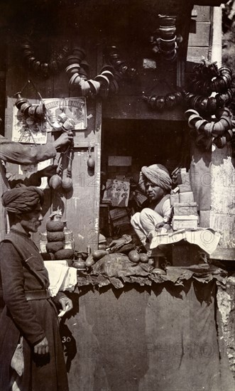 Roadside stall at Mussoorie bazaar. An Indian man sits hunched up on a roadside stall in a bazaar at Mussoorie. He appears to be a haberdasher, selling items including scissors and dress-making patterns. Mussoorie, India, circa 1890. Mussoorie, Uttaranchal, India, Southern Asia, Asia.