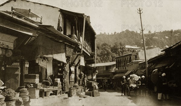 Bazaar at Mussoorie, India. Ramshackle buildings line a winding road at a bazaar in Mussoorie. A European woman visits the market stalls, pulled along in a rickshaw by uniformed Indian servants. Mussoorie, India, circa 1890. Mussoorie, Uttaranchal, India, Southern Asia, Asia.