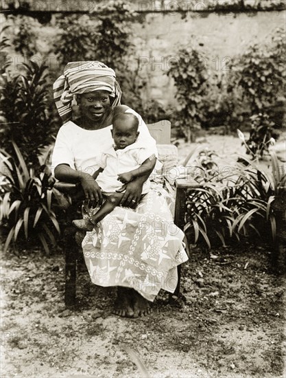 Mother and child, Nigeria. Portrait of a seated African woman holding a baby on her knee. She wears traditional clothing including a head scarf and a patterned wrap-around skirt. Nigeria, circa 1925. Nigeria, Western Africa, Africa.