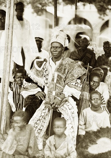 Nigerian chief, Badagry. An unidentified chief, perhaps the Akran of Badagry, is seated wearing elaborate embroidered robes with a high-sided and tasselled cap. He holds a staff of office and is surrounded by children sitting on the ground, one of whom holds a fan edged with feathers. Badagry, Nigeria, 1927. Badagry, Lagos, Nigeria, Western Africa, Africa.