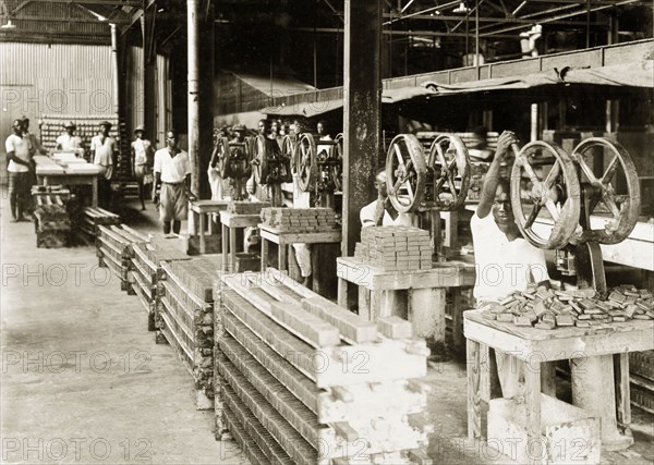 Soap company stamping room. A row of African factory workers stamp bars of soap with hand-operated presses at the West African Soap Company in Apapa. Lagos, Nigeria, circa 1925. Lagos, Lagos, Nigeria, Western Africa, Africa.