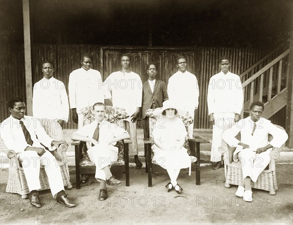 The Baxendales with Miller Brothers' staff. Mr and Mrs Baxendale (centre) pose for the camera with African employees of Miller Brothers & Co. The six young African men behind them are dressed mostly in white, possibly house servants or office staff. Two African men in formal Western dress sit on either side of the Baxendales in cane armchairs. Badagry, Nigeria, circa 1924. Badagry, Lagos, Nigeria, Western Africa, Africa.