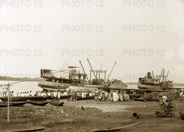 Boats at a Badagry wharf. Two steamers, labelled in manuscript as 'Ife' and 'Ila', are moored at a wharf shared by Miller Brothers & Co. and the African Oil Nuts Company. People crowd around both boats. Badagry, Nigeria, circa, 1925. Badagry, Lagos, Nigeria, Western Africa, Africa.