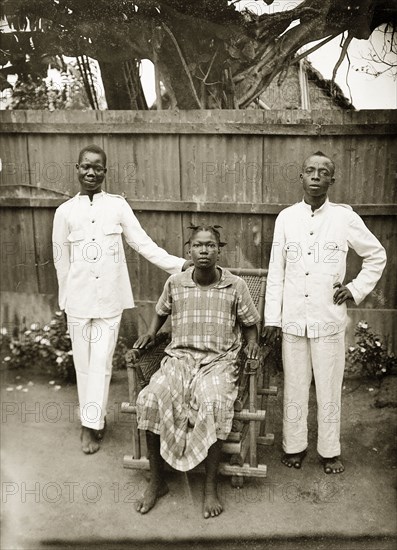 Portrait of domestic servants. Portrait of two male domestic servants and a woman in a garden. James and Godfrey both wear white uniforms, Godfrey's wife (seated) wears a checked knee-length dress. All are barefoot. Badagry, Nigeria, circa 1928. Badagry, Lagos, Nigeria, Western Africa, Africa.